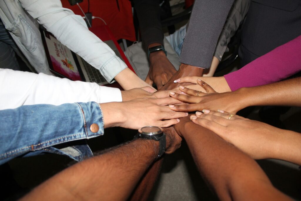 A photograph showing the forearms and hands of several different people with different skin tones, different jewelry, and different shirt sleeve lengths and colors. Everyone's hands are piled on top of each other in a circle as if they were standing in a circle and each put one hand in as a signal of a team.
